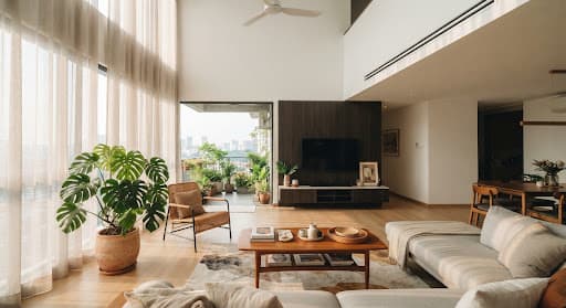 Sunlit high-ceiling contemporary Malaysian living room with light wood floors, sheer curtains filtering afternoon warm sunlight, indoor potted tropical monstera plants, and a minimalist flush architectural ducted AC grille integrated cleanly into the high white ceiling.